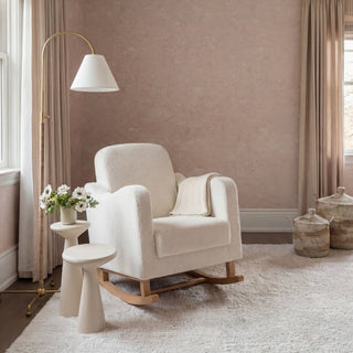 White rocking chair with a blanket, a small table with flowers, a floor lamp, and woven baskets in a blush-colored room.