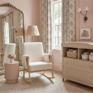 Rocking chair next to pink side table with flowers, in a room featuring light curtains and a dresser with baskets.