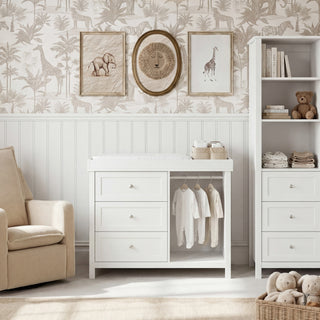 Changing table with three drawers, hanging white baby clothes, beige armchair, and shelves with toys in nursery room.