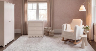 White rocking chair with a throw blanket, next to a small table with flowers, and a dresser under a window.